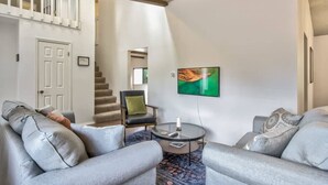 Un salon avec des canapés gris, une table basse, un téléviseur et un escalier menant à l’étage supérieur.
