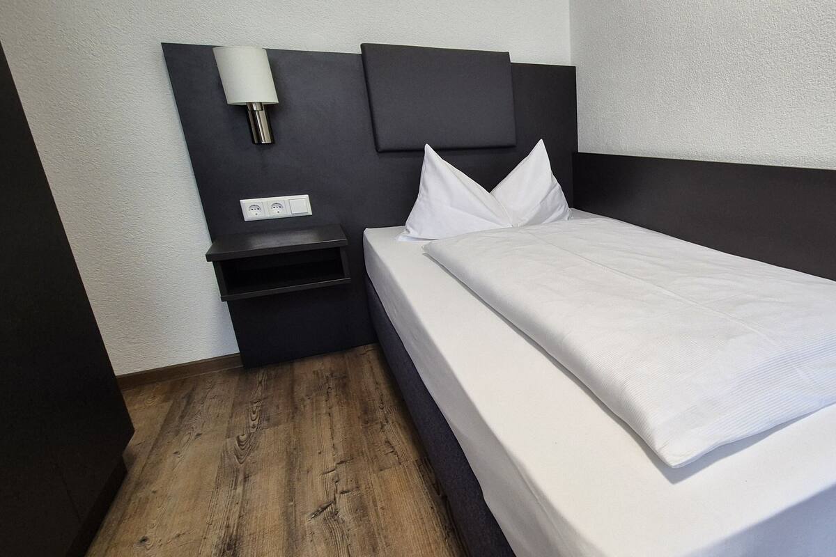 A single bed with white bedding, a dark headboard, and a bedside table with a lamp.