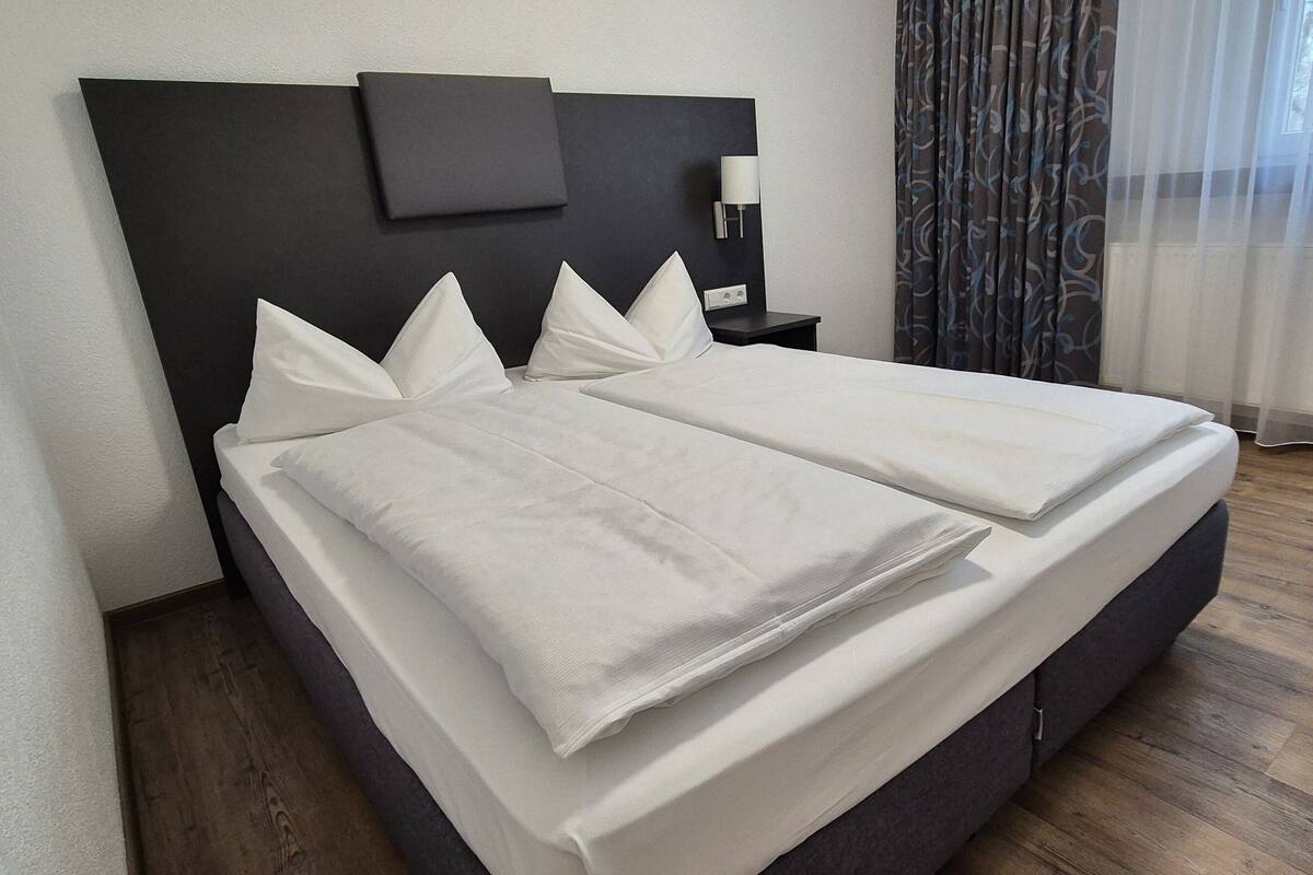 A double bed with white linens, a dark headboard, a bedside table with a lamp, and a chair.