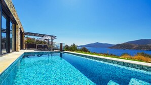 A swimming pool with clear blue water, surrounded by a modern building with large windows and a view of the sea and mountains. - Villa Nana, A Modern 1 Bedroom Property in Komurluk (Antalya)