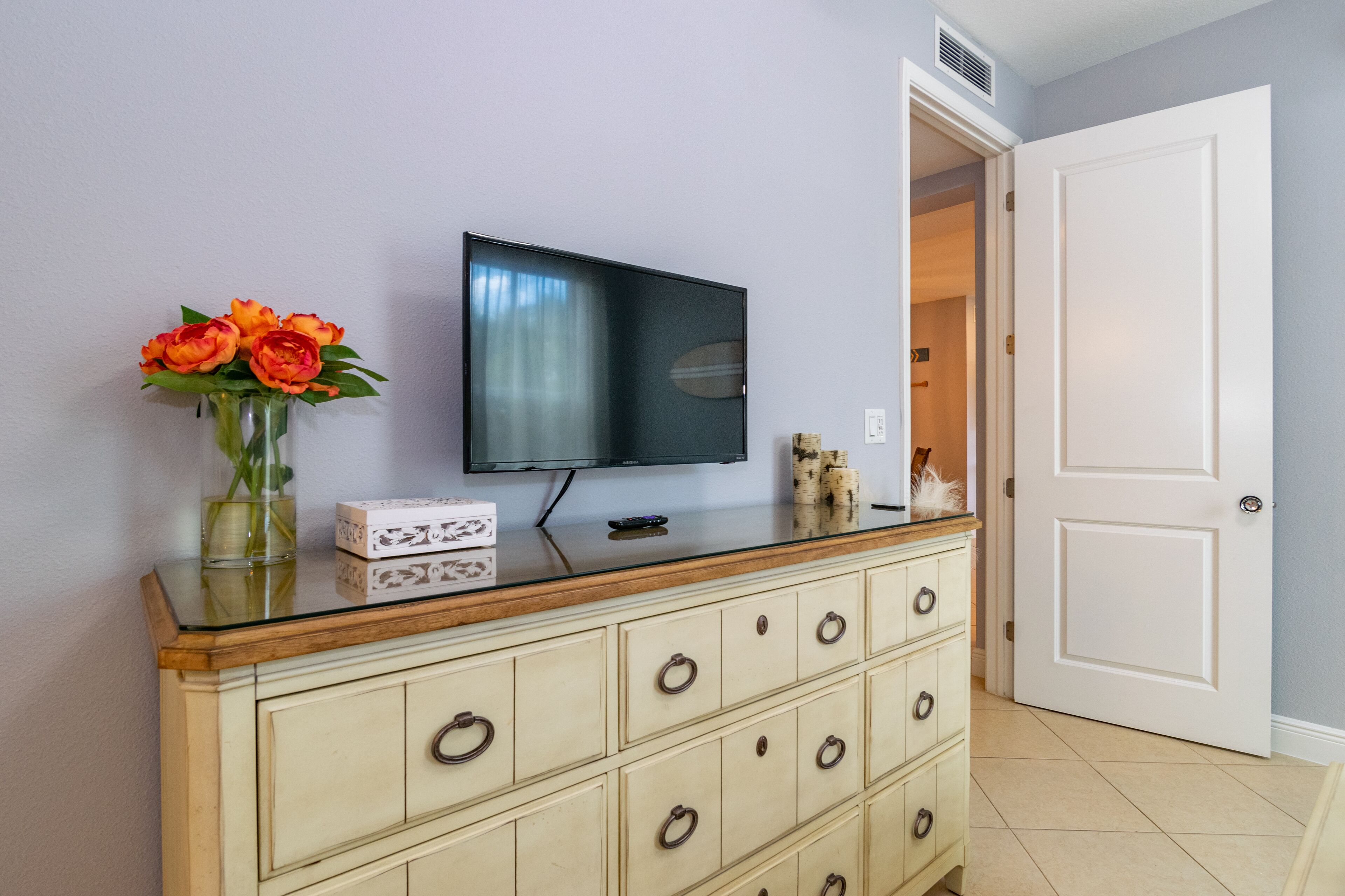 Quarto com televisão sobre um armário de madeira, um vaso com flores e uma porta branca.
