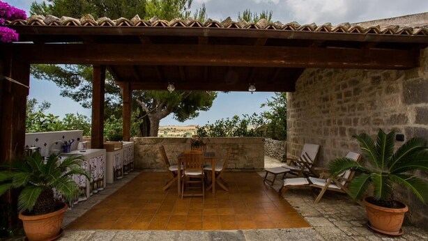 Un patio techado al aire libre con suelo de piedra, mobiliario de madera y plantas en macetas.