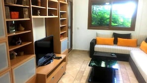 Un soggiorno con televisore, scaffali in legno, un divano con cuscini arancioni e una finestra vista verde.