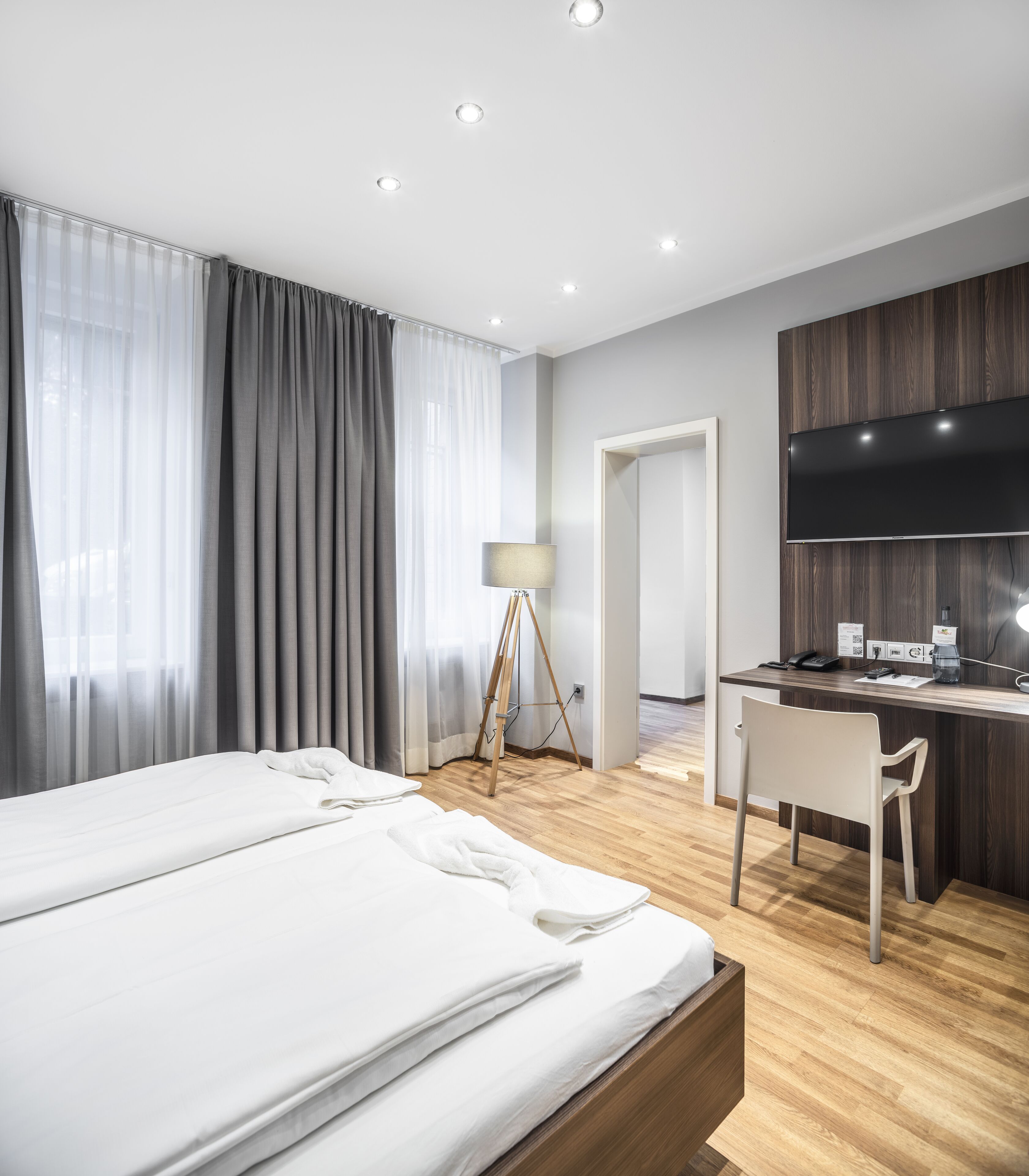 A modern hotel room with a large bed, a desk, a chair, a lamp, and a flat-screen TV.