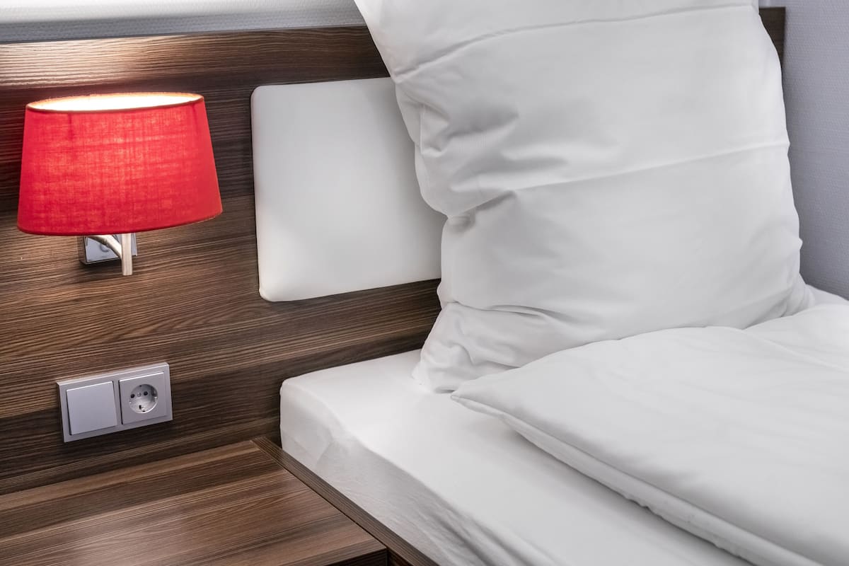 Una cama bien hecha con sábanas blancas y una lámpara roja sobre una mesita de noche de madera.