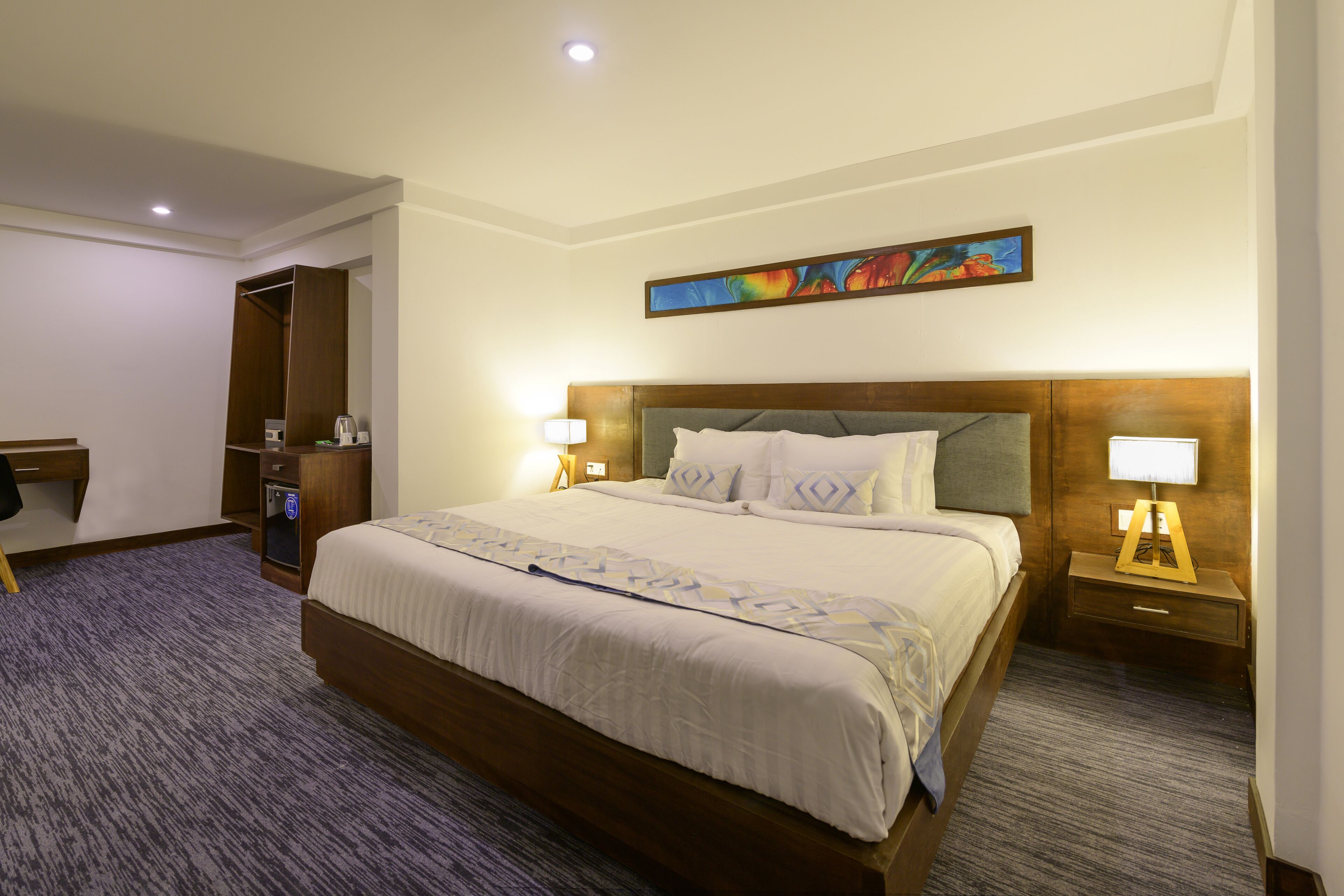 A hotel room with a large bed, a wardrobe, a desk, and a painting on the wall.