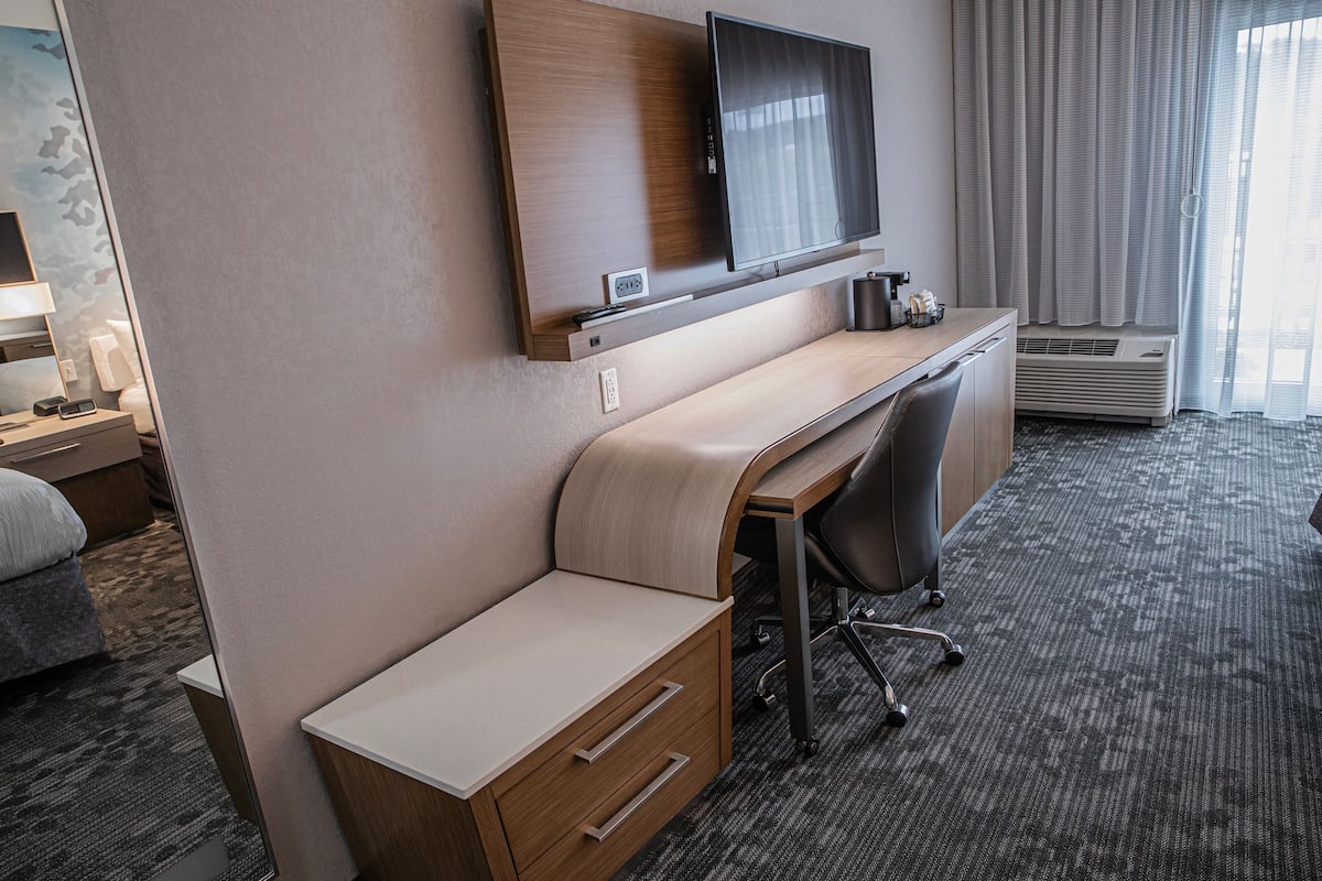 A hotel room with a flat-screen TV mounted on a wooden panel, a desk with a chair, a small bedside table with a lamp, and a large window with curtains.
