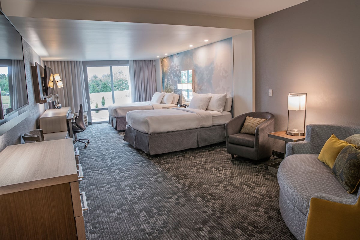 A hotel room with a large bed, a desk, a sofa, and a chair. There is a window with curtains and a view of trees.