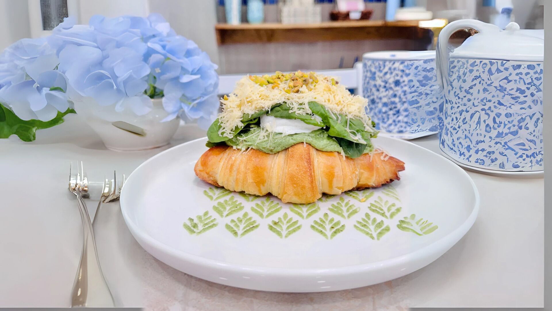 A croissant with spinach and cheese on a white plate, accompanied by a blue and white patterned cup and a vase of blue flowers.