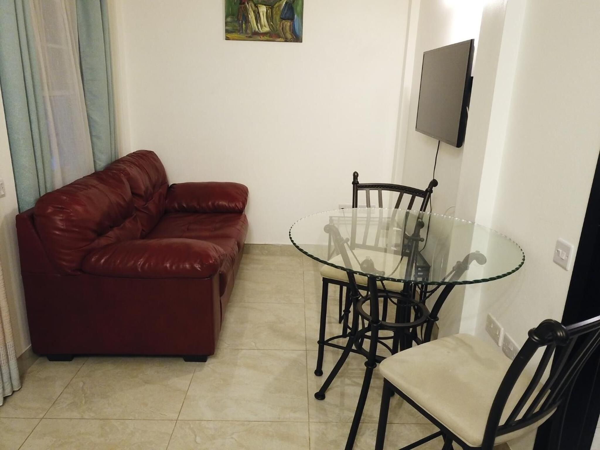 A living room with a red leather sofa, a glass table with four chairs, and a flat-screen TV mounted on the wall.