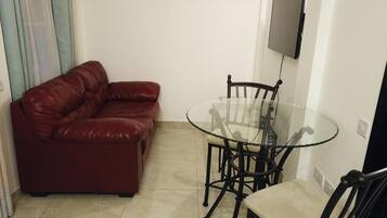 A living room with a red leather sofa, a glass table with four chairs, and a flat-screen TV mounted on the wall.
