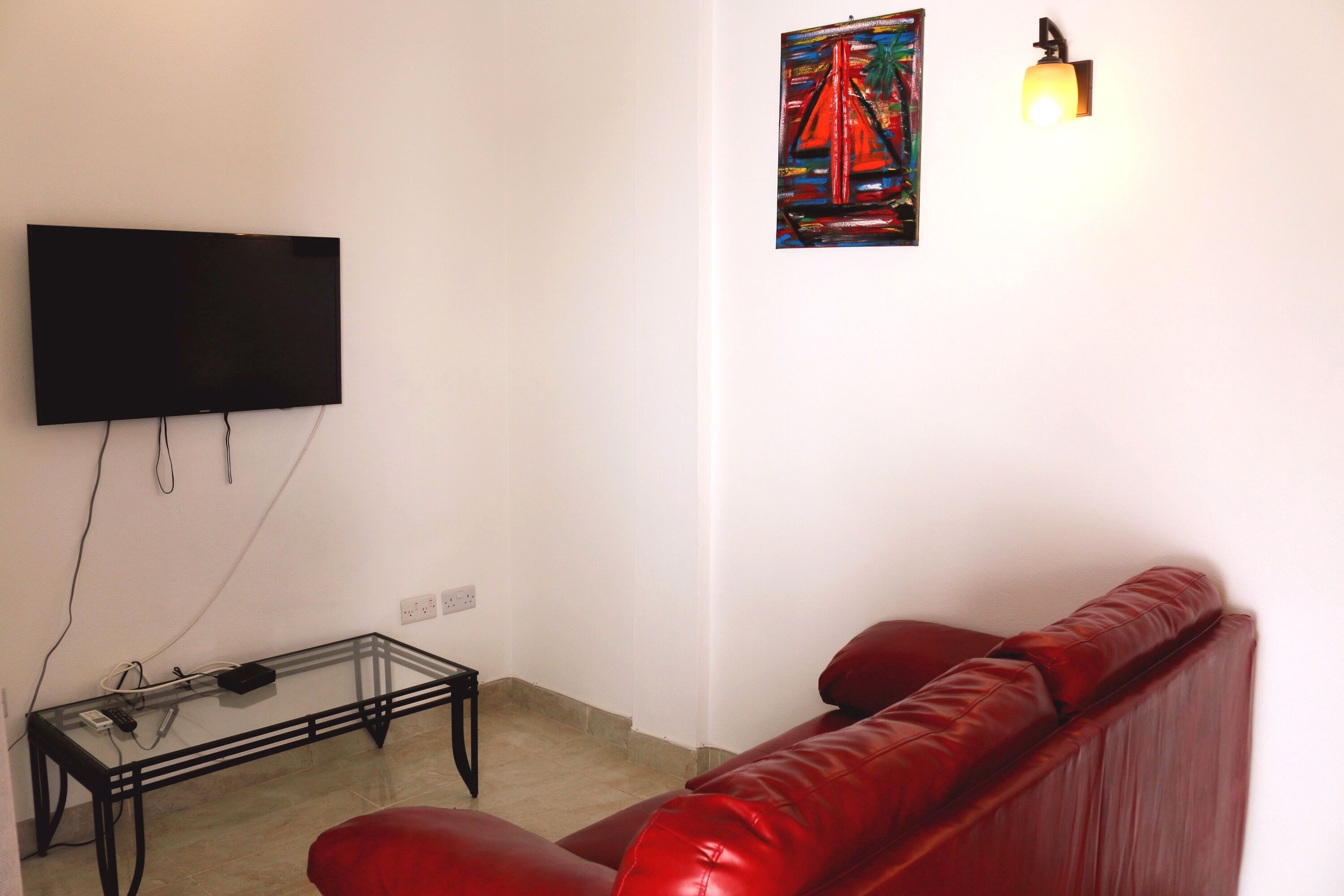 Een woonkamer met een flatscreen televisie aan de muur, een glazen salontafel met elektronische apparaten, een rode leren bank en een schilderij van een brug.