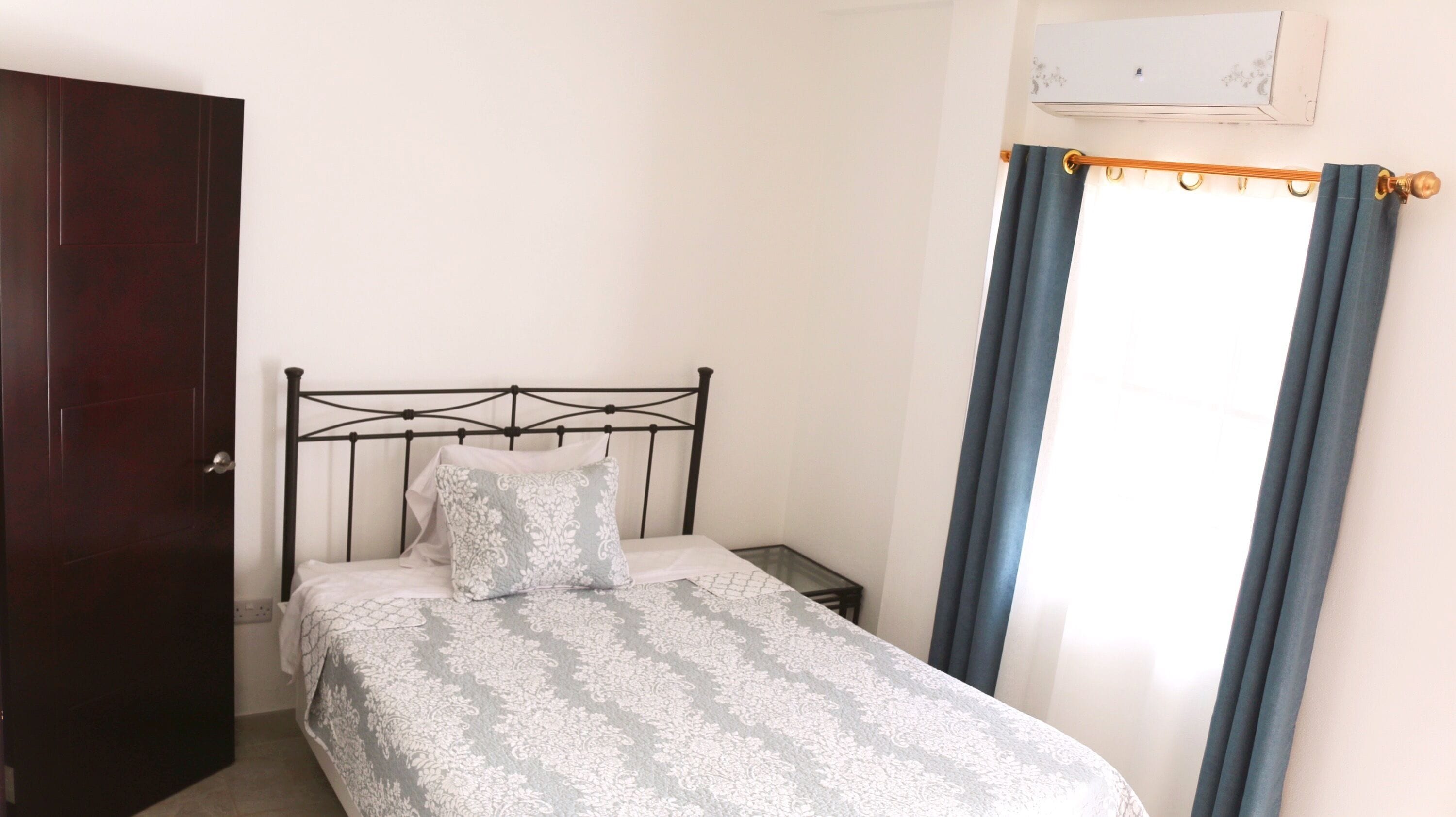 A bedroom with a bed, a nightstand, a wooden door, and a window with curtains.