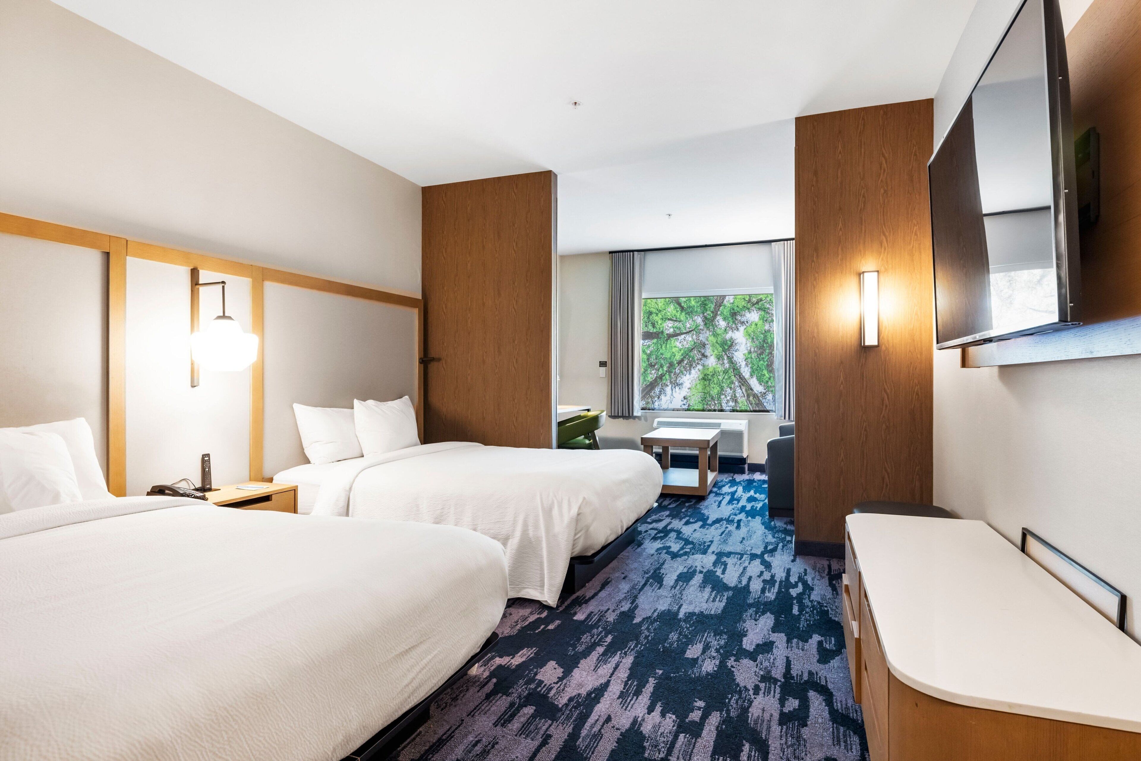 A hotel room with two beds, a desk, a chair, a TV, and a window with a view of trees.