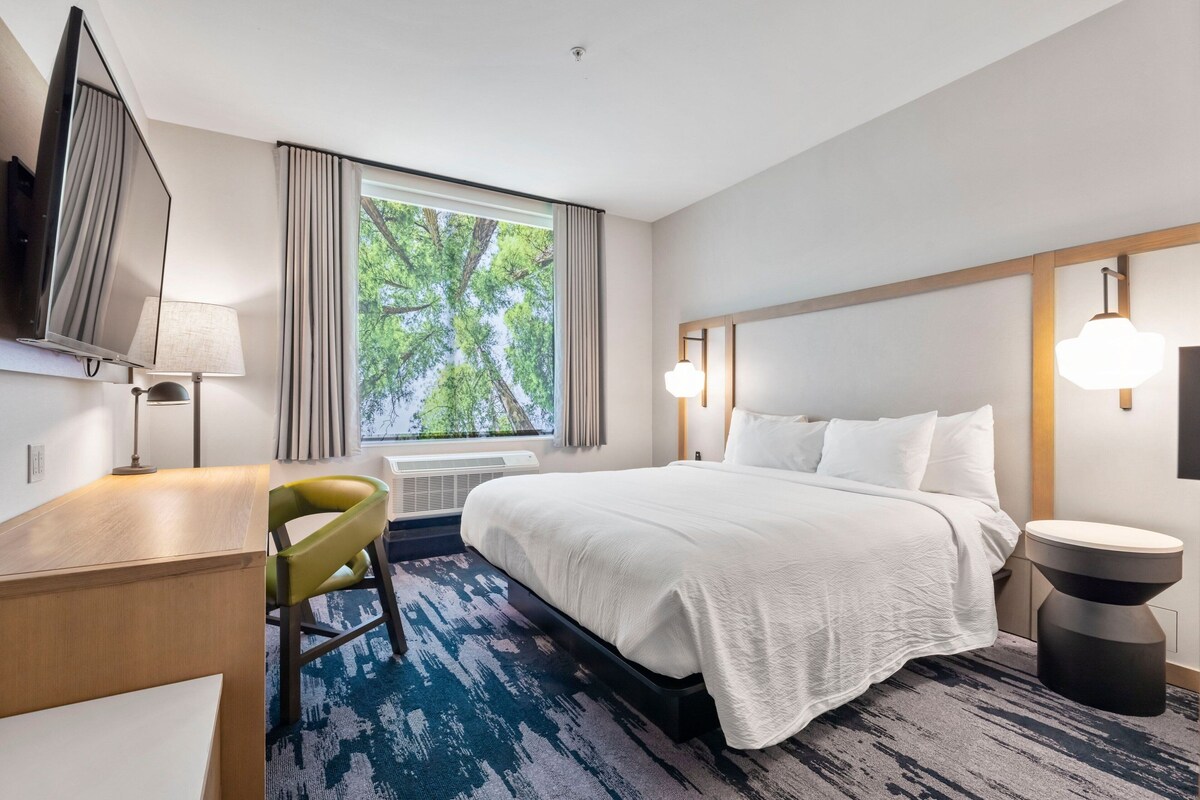 Habitación de hotel con cama, escritorio, silla, televisor, ventana con cortinas y vistas a árboles.