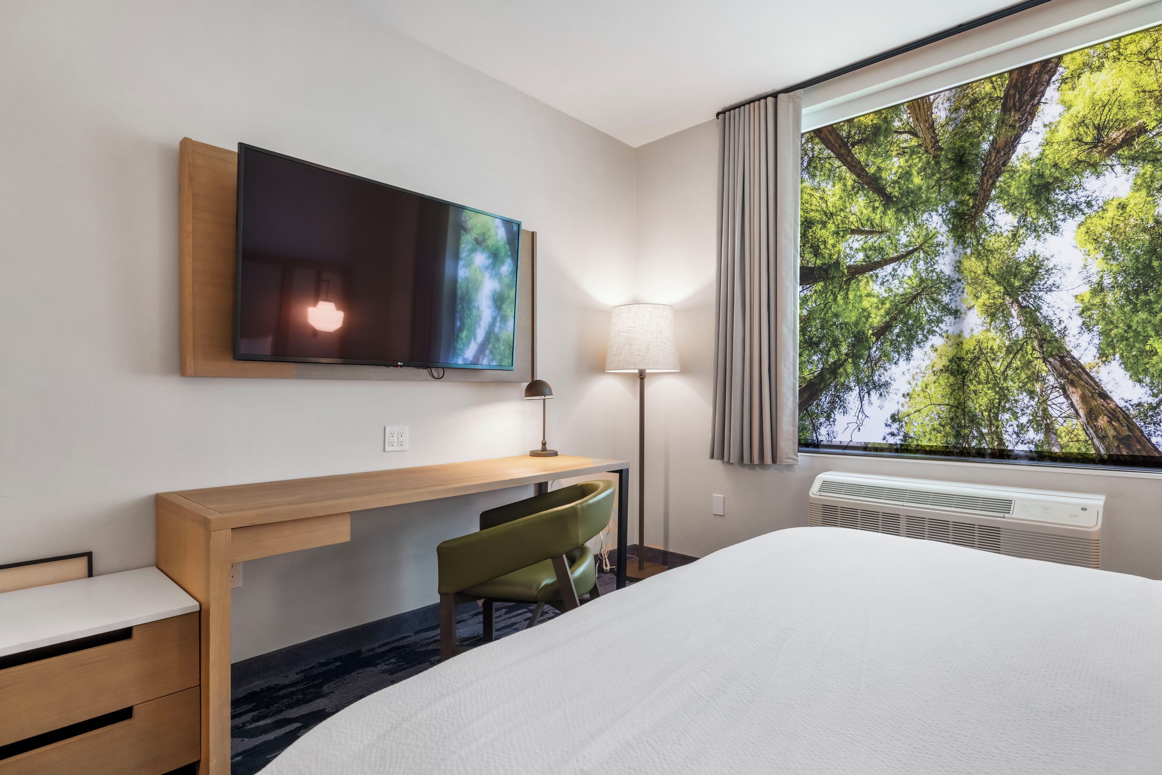A hotel room with a bed, a desk, a chair, a TV mounted on the wall, a lamp, and a window with a view of trees.