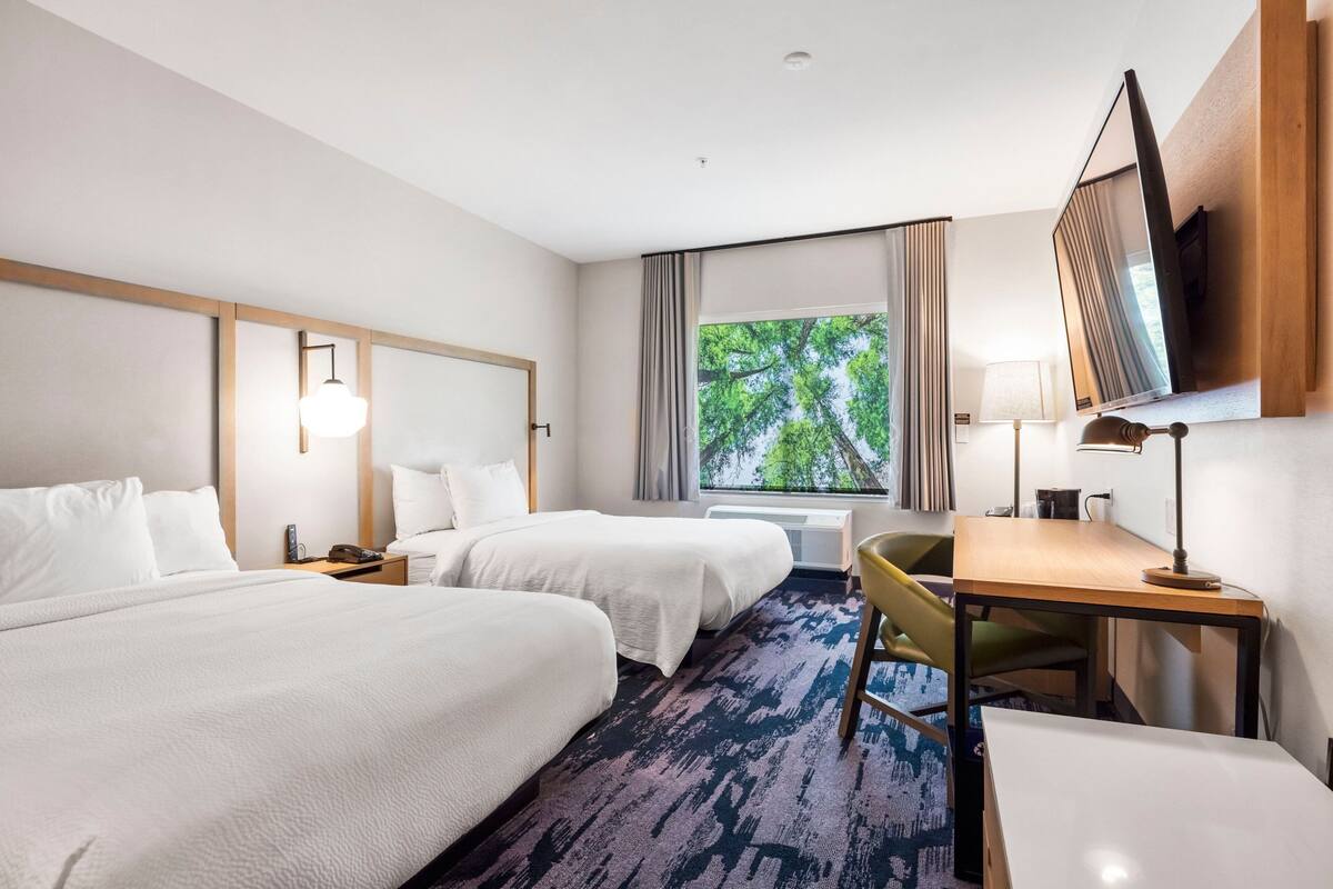 Habitación de hotel con dos camas, un escritorio, una silla, un televisor y una ventana con vista a árboles.