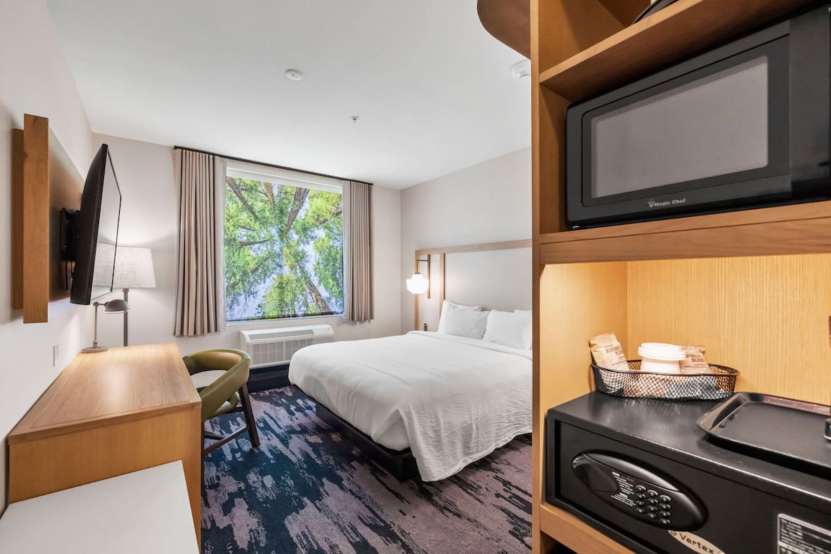 Habitación de hotel con cama, escritorio, televisor, microondas y ventana con vista a árboles.