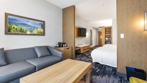 Habitación de hotel con sofá, una mesa de madera, cama, escritorio, televisor y una ventana grande con vistas a un bosque.