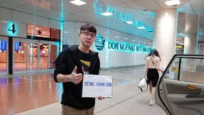 En person holder et skilt med tekst, stående i et moderne indendørsområde med glasdøre og et skilt, der står 'DON MUEANG INTERNATIONAL'.