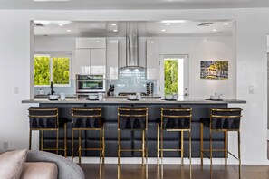 A modern kitchen with a central island, bar stools, and a view of greenery outside.