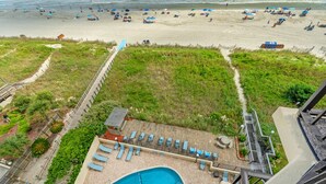 A rooftop pool area with lounge chairs and a small pool, overlooking a beach with umbrellas and people.