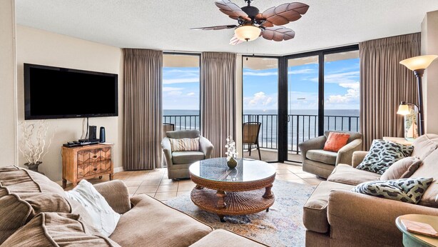 A spacious living room with a large sofa, a coffee table, and a television. There is a balcony with a view of the ocean.