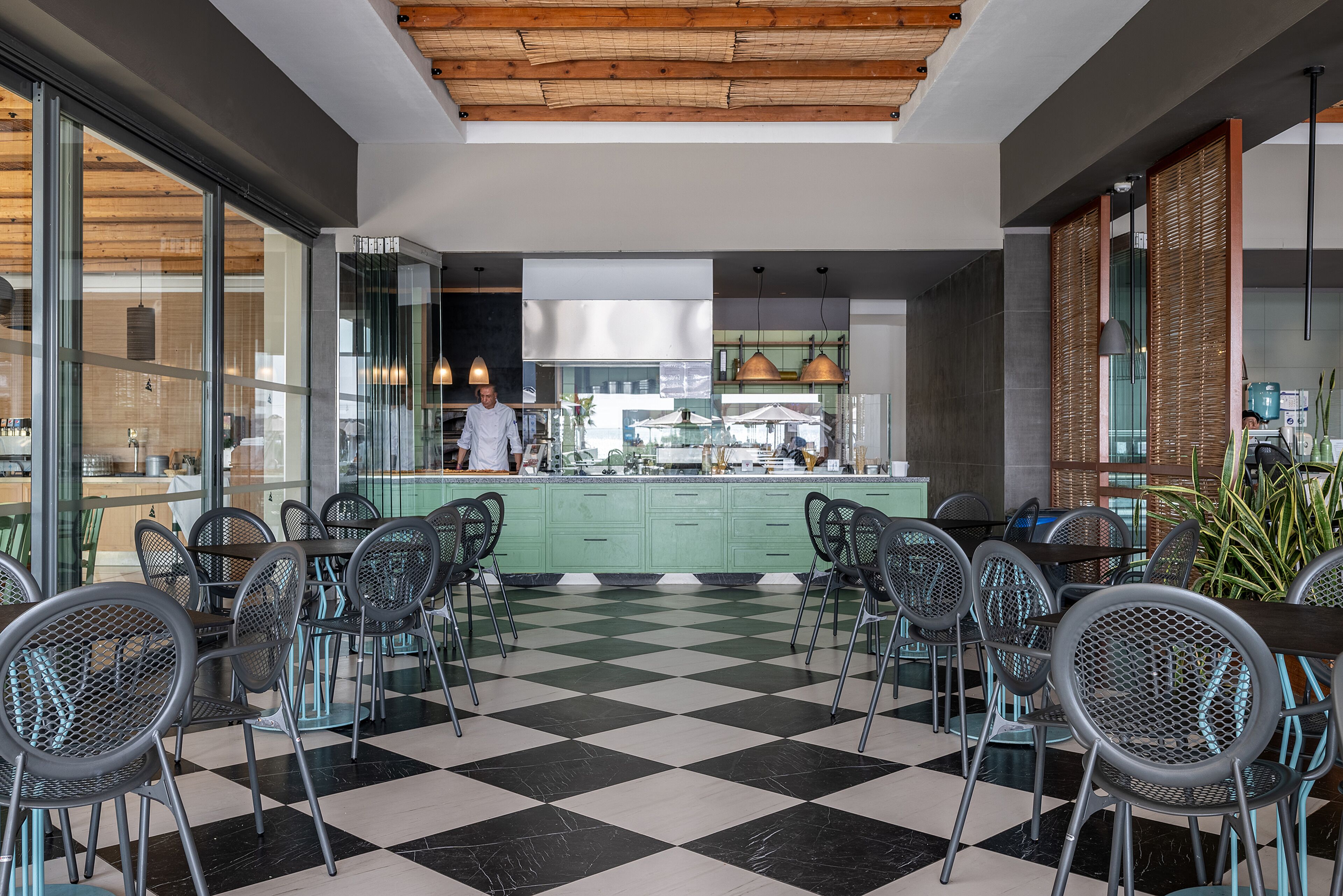 Un restaurante con piso a cuadros, sillas metálicas y un chef preparando comida.