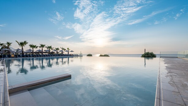 Una piscina infinita tranquila con agua cristalina que refleja el cielo, rodeada de palmeras y sillones de descanso.