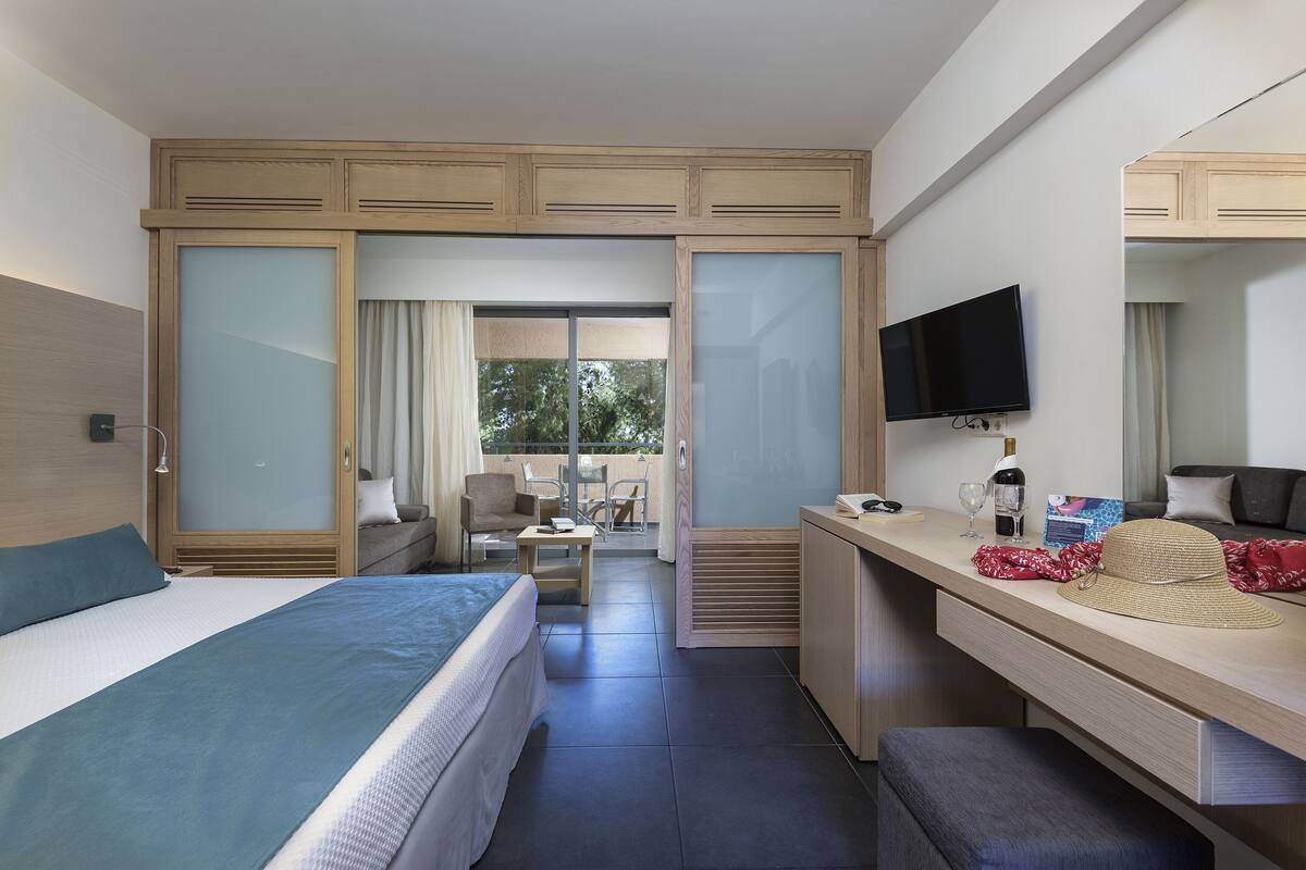 Hotelkamer met een bed, bureau, televisie en balkon met uitzicht op bomen.