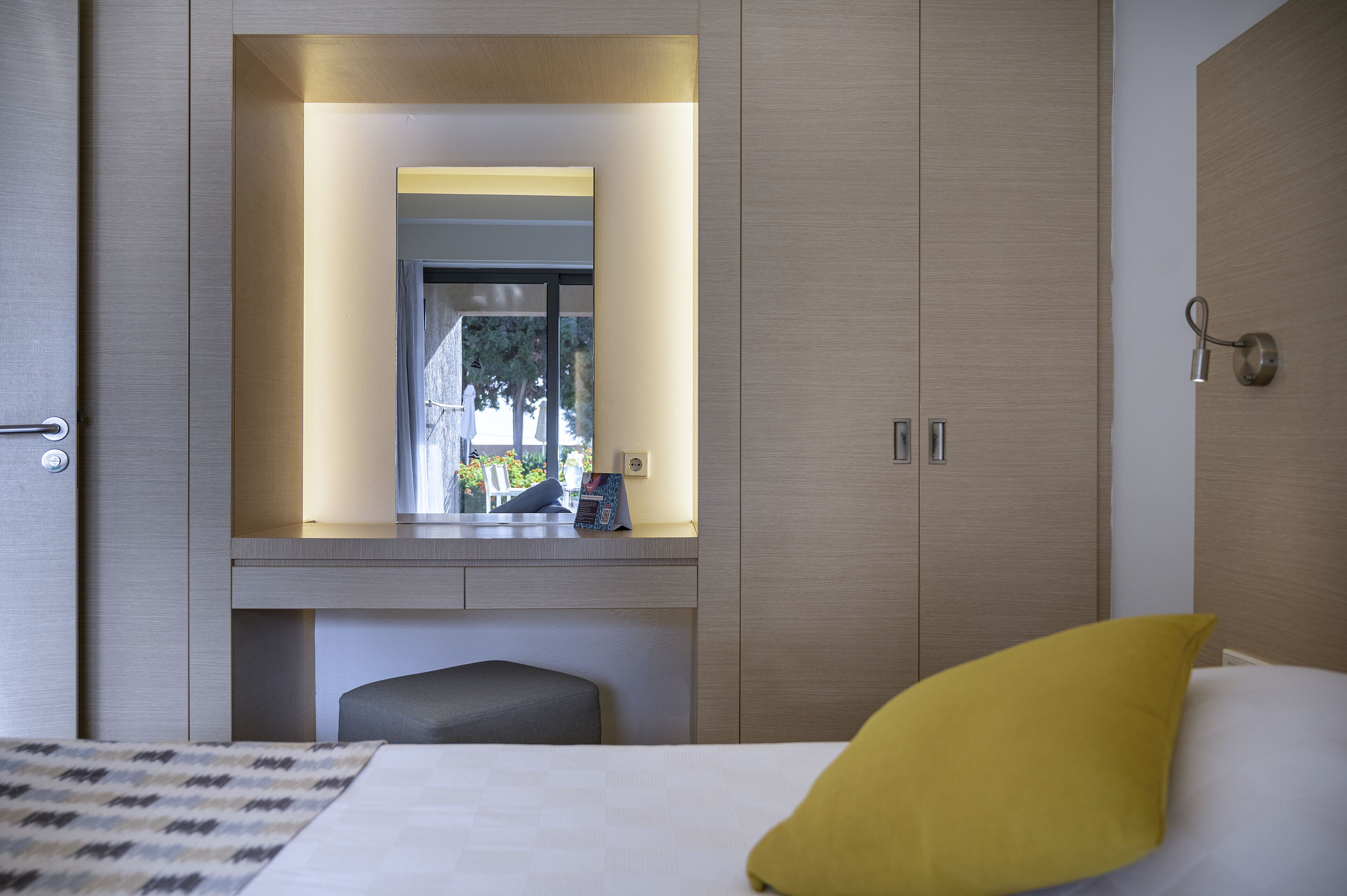 Une chambre d’hôtel moderne avec un grand miroir, une table de chevet, un lit avec un oreiller jaune et une vue sur l’extérieur par la fenêtre.