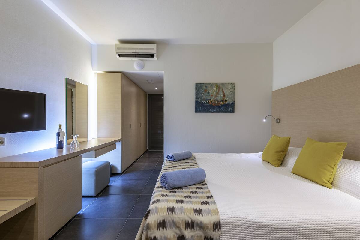 Een moderne hotelkamer met een flatscreen televisie, een bureau met een fles en glas, een bed met een bedsprei met patroon en twee kussens, en een schilderij aan de muur.