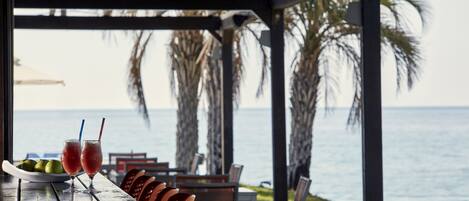 A covered outdoor dining area with a view of the sea, featuring a table with two drinks and a plate of fruit, surrounded by chairs.