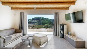 Ein modernes Wohnzimmer mit einer Couch, einem Couchtisch, einem abgedeckten Hocker, einem an der Wand montierten Fernseher und Blick auf eine grüne Landschaft durch große Fenster.