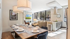 A modern dining area with a wooden table set for six, a sofa, and a kitchen in the background.