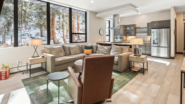 A modern living room with a sectional sofa, a coffee table, and a view of a forest through large windows.