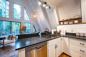Enjoy a meal with a view of the surrounding forest from this kitchen, featuring a large window, dishwasher, and comfortable seating area.