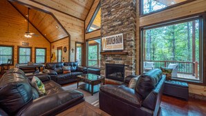A cabin with a stone fireplace, leather sofas, and large windows offering forest views. - # Kick Back Shack  ## Luxury Broken Bow Cabin with Hot Tub, Pool Table, EV Charg (Broken Bow)