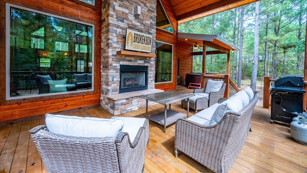 A cabin with a stone fireplace, wicker furniture, and a large window overlooking the forest. - # Kick Back Shack  ## Luxury Broken Bow Cabin with Hot Tub, Pool Table, EV Charg (Broken Bow)