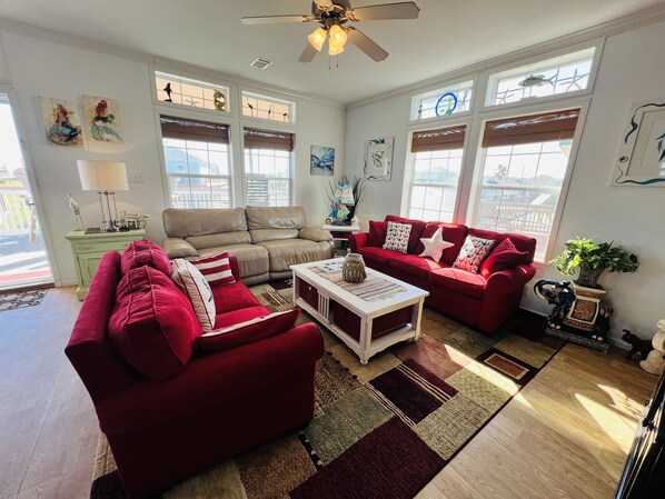 A spacious living room with a beige sofa, red armchairs, and a white coffee table. Large windows provide ample natural light, and the room features a ceiling fan and decorative wall art. - Home with beach view from front porch, only two minute walk to beach. Very Nice! (Crystal Beach)