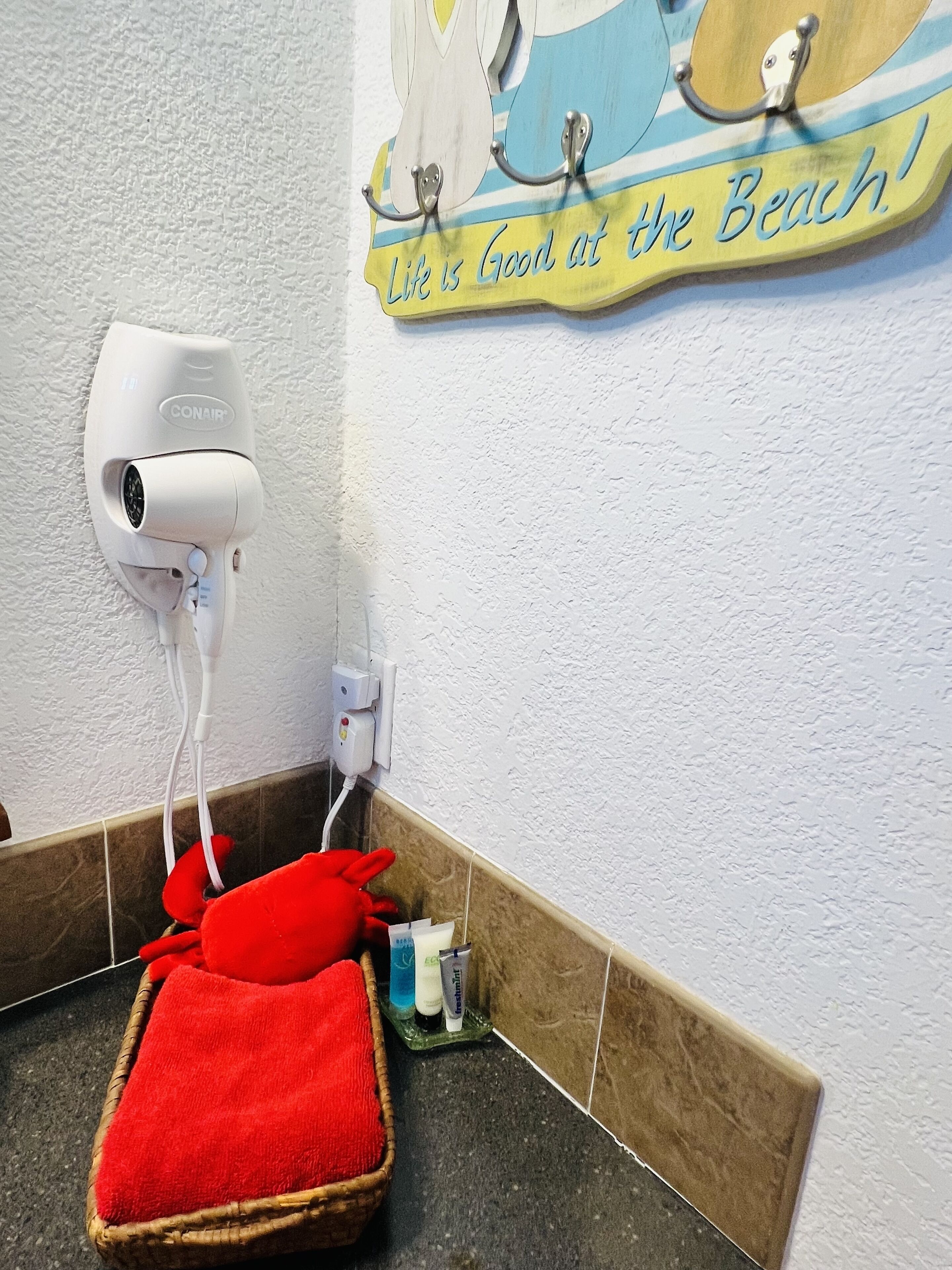 A bathroom with a hairdryer mounted on the wall, a towel rack with a towel, and a sign that reads "Life is Good at the Beach!"