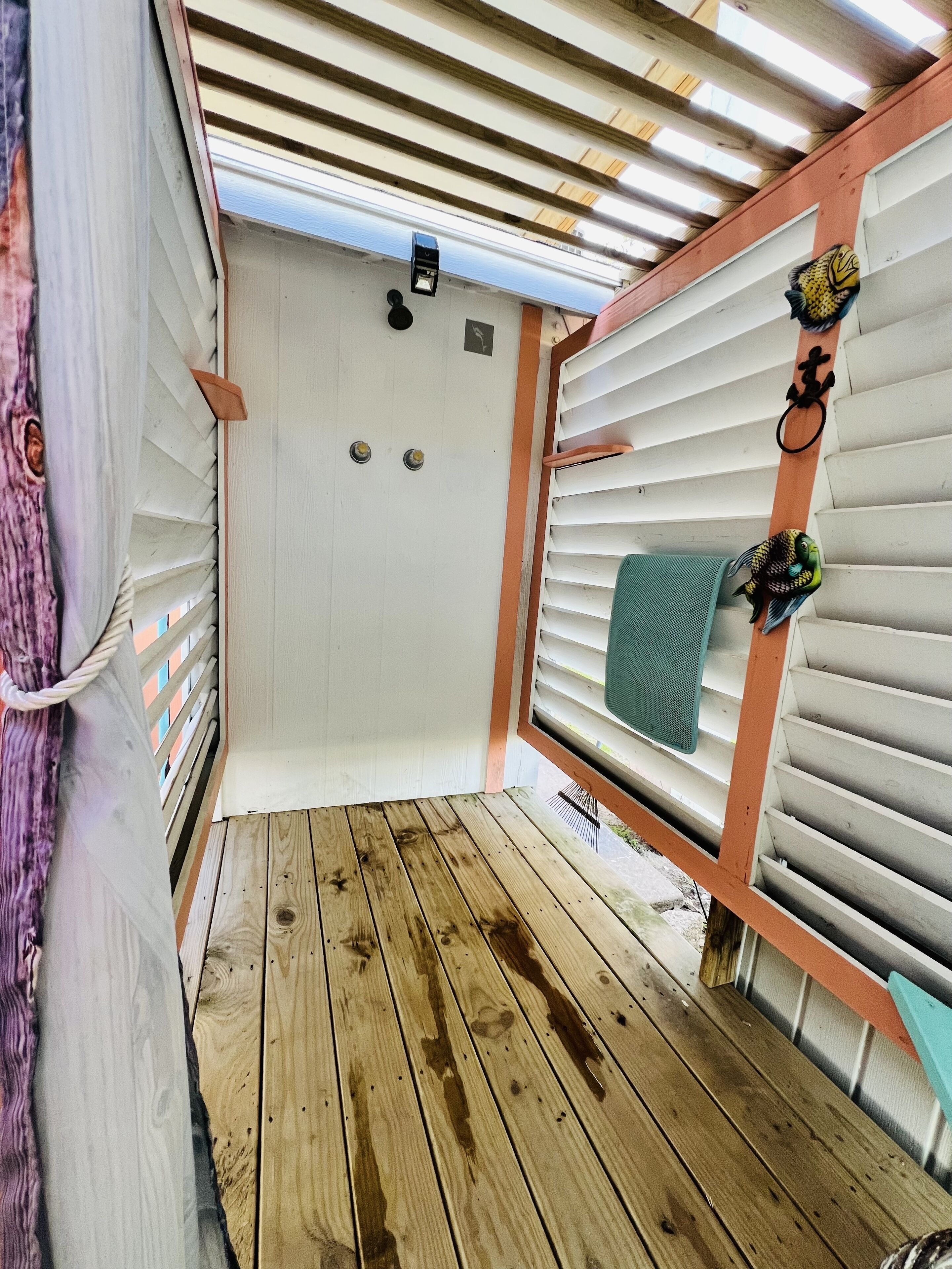A covered outdoor area with a wooden deck, white walls with horizontal slats, and a small window with a towel hanging on the right side.
