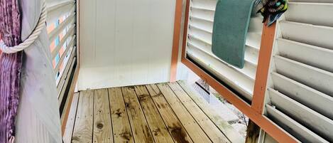 A covered outdoor area with a wooden deck, white walls with horizontal slats, and a small window with a towel hanging on the right side.