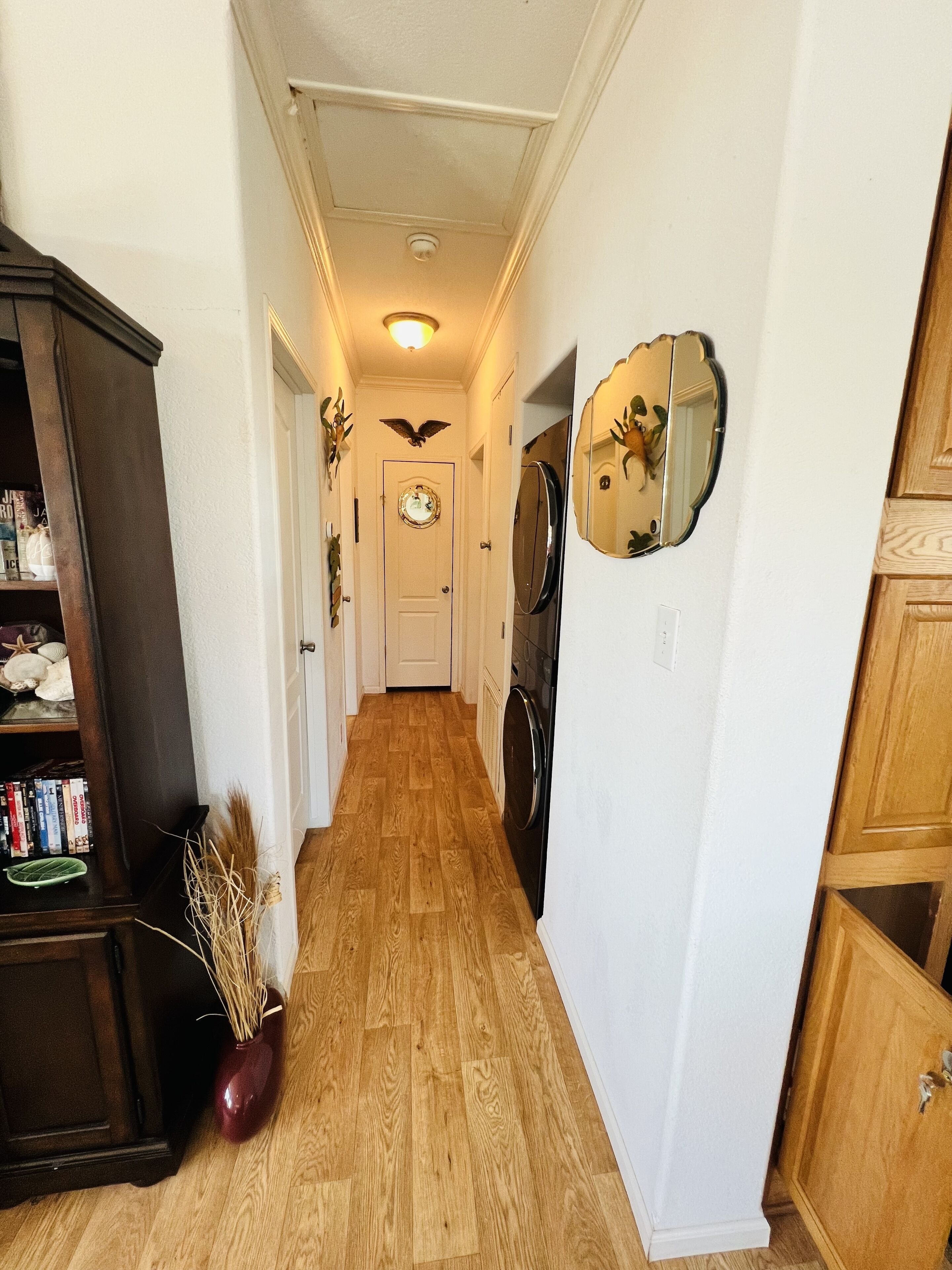 A narrow hallway with wooden flooring, a dark wooden cabinet on the left, a vase with dried plants, and a mirror on the right wall.