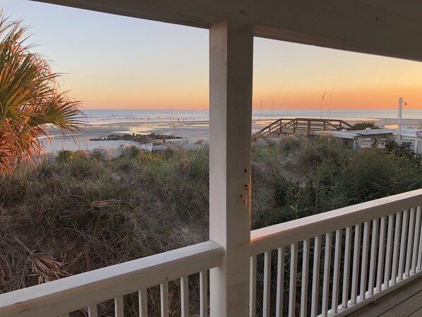 Uitzicht op een strand vanaf een overdekt terras met houten balustrade, een rotsachtig strand en een palmboom.