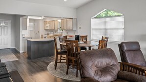 Een ruime woonkamer met een grote bruine leren bank, een eethoek met houten stoelen, en een keuken op de achtergrond.