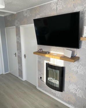 Una habitación con un televisor de pantalla plana montado en la pared, un estante de madera debajo y una chimenea con un faldón blanco.