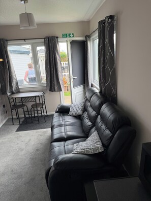 A room with a black leather sofa, a dining table with chairs, a window with curtains, and a fire exit sign. - Chalet 91A South Shore Bridlington, Sleeps 4 (Bridlington)