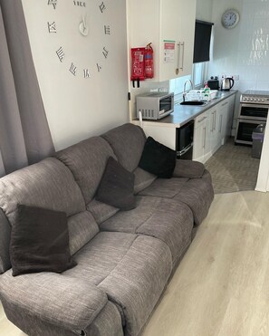 Un salon moderne avec un canapé gris, un coin cuisine équipé d’un micro-ondes, d’un grille-pain et d’un four, et une horloge murale.