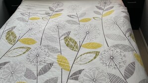 A neatly made bed with a floral-patterned bedspread, a bedside lamp, and a window with sheer curtains. - Chalet 91A South Shore Bridlington (Bridlington)